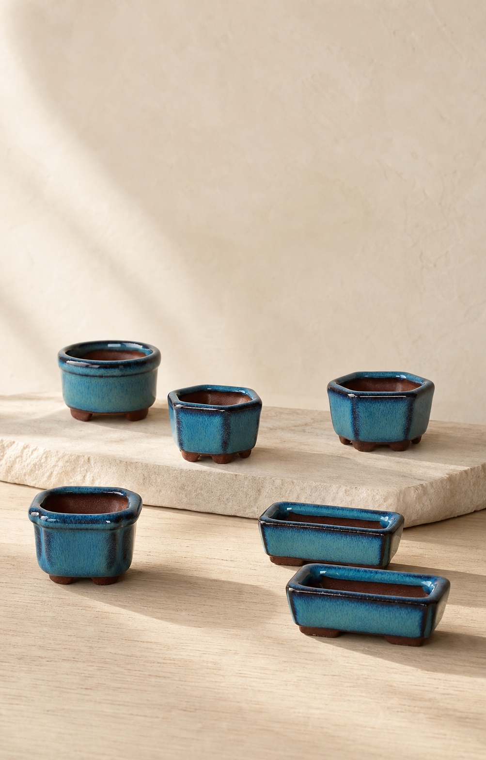 A curated collection of fingertip-sized blue-green glazed bonsai pots on warm stone and pale wood