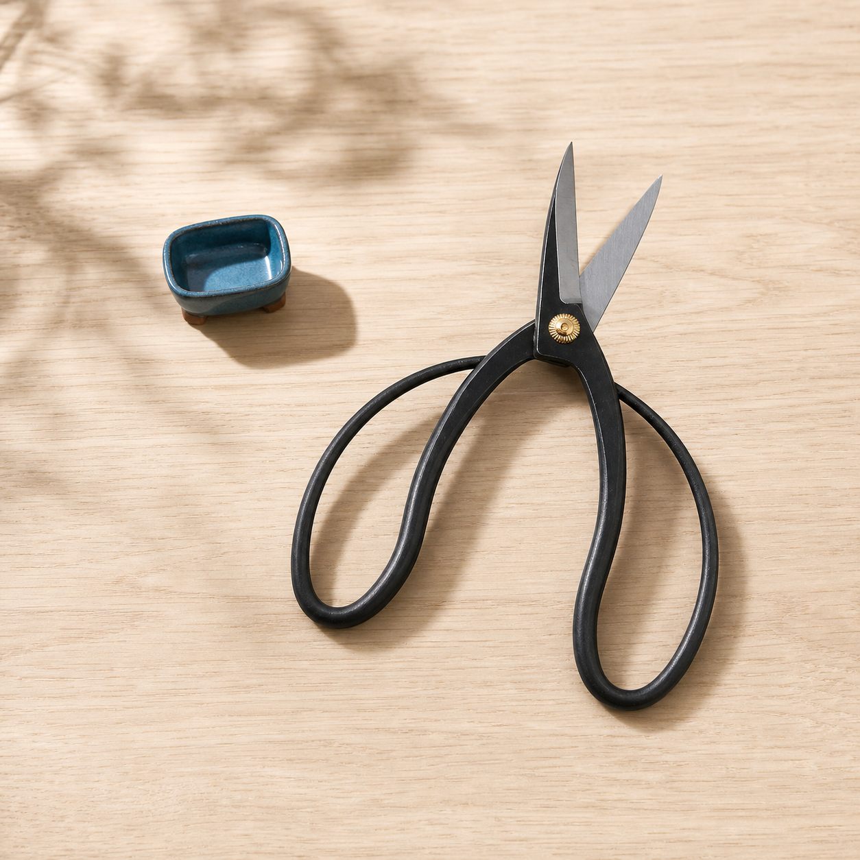 Black loop-handled 180 mm Wazakura bonsai scissors with silver blades and a brass pivot beside a fingertip-sized blue bonsai pot